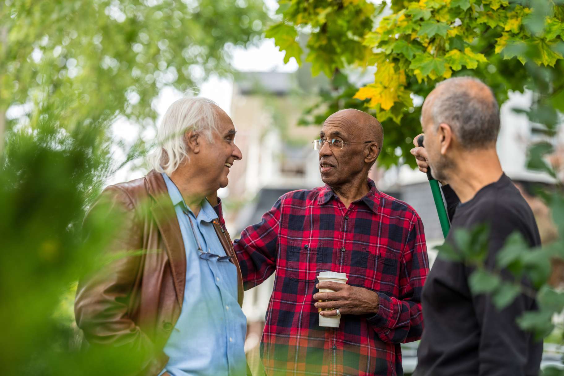 Foto på tre män som står och samtalar utom hus. En av dem håller i en mugg och en i något som ser ut att vara en kratta. 